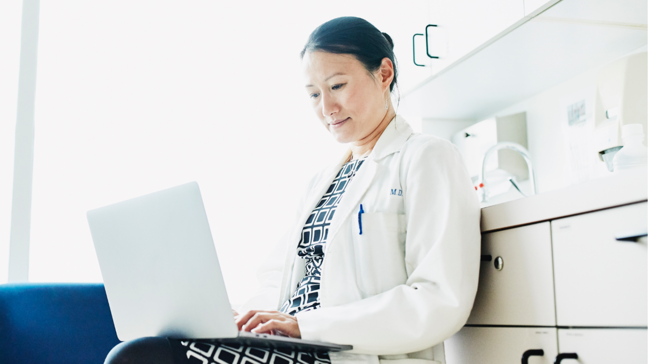Female healthcare provider in a white medical coat in an office working on a laptop