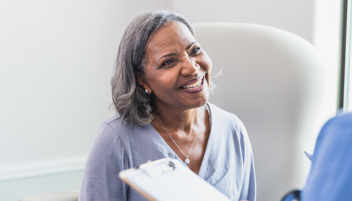 Smiling middle aged woman with graying hair speaking to a healthcare provider holding a clipboard